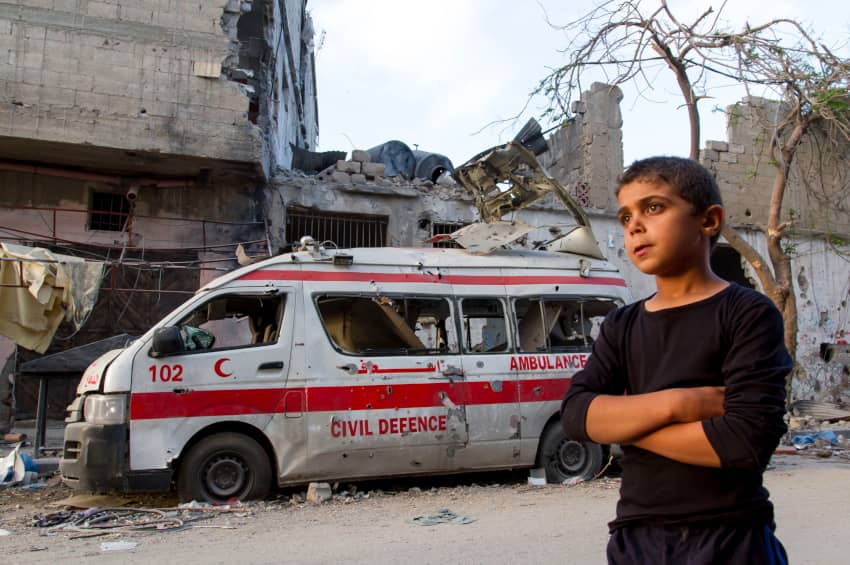 Niño palestino de pie junto a una ambulancia destruida y edificios en ruinas tras bombardeos israelíes