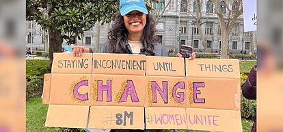 Activista feminista con un cartel en inglés que reclama cambios frente a la violencia contra las mujeres durante una protesta en la calle.