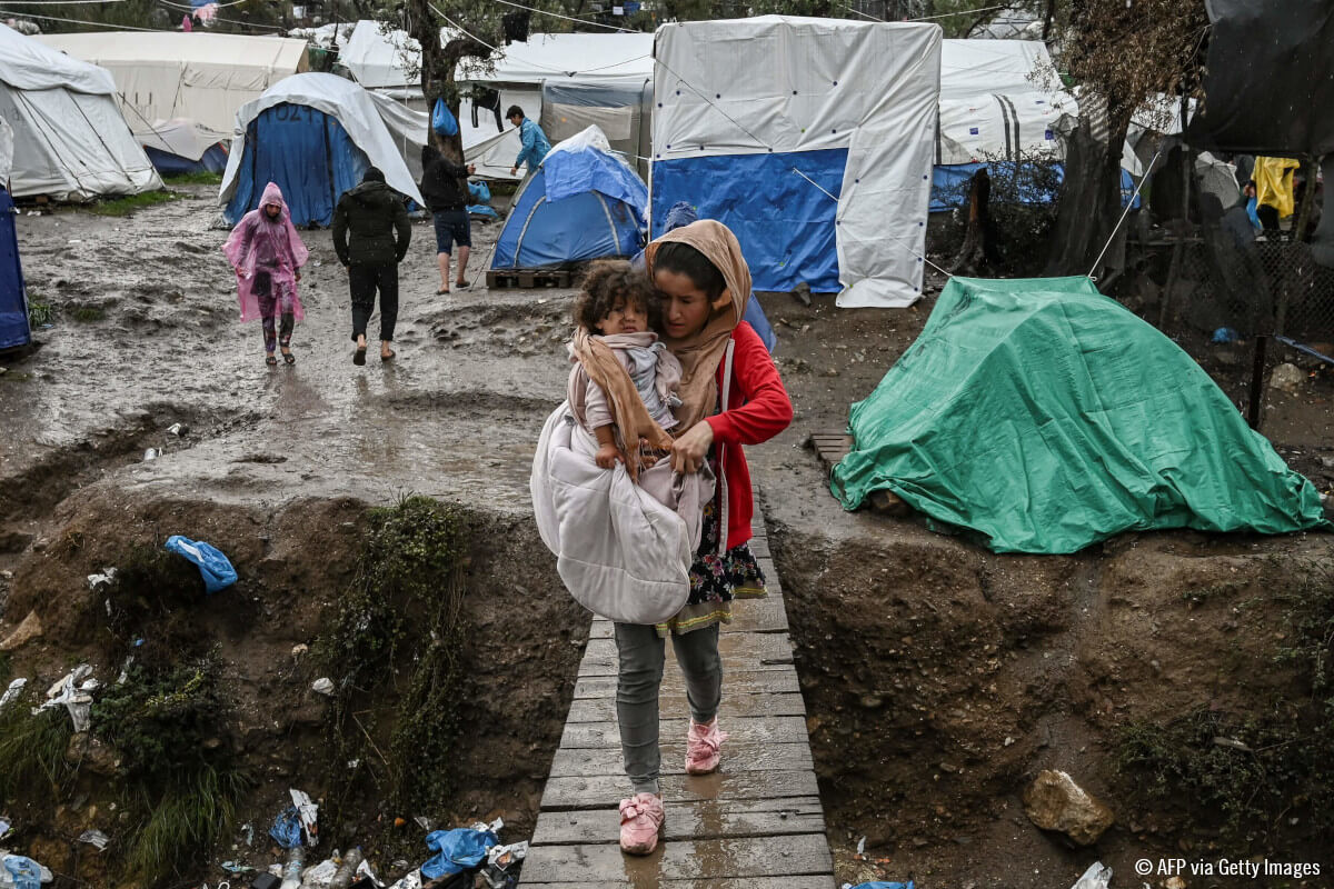 Una mujer refugiada cargando a una niña. Al fondo, tiendas de campaña