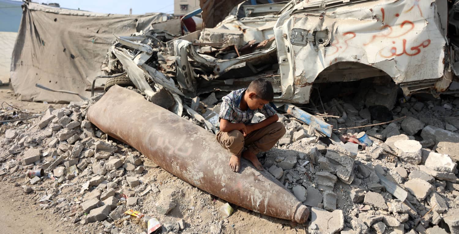 Niño palestino sentado sobre un proyectil sin explotar en medio de escombros y restos de vehículos destruidos en Gaza.
