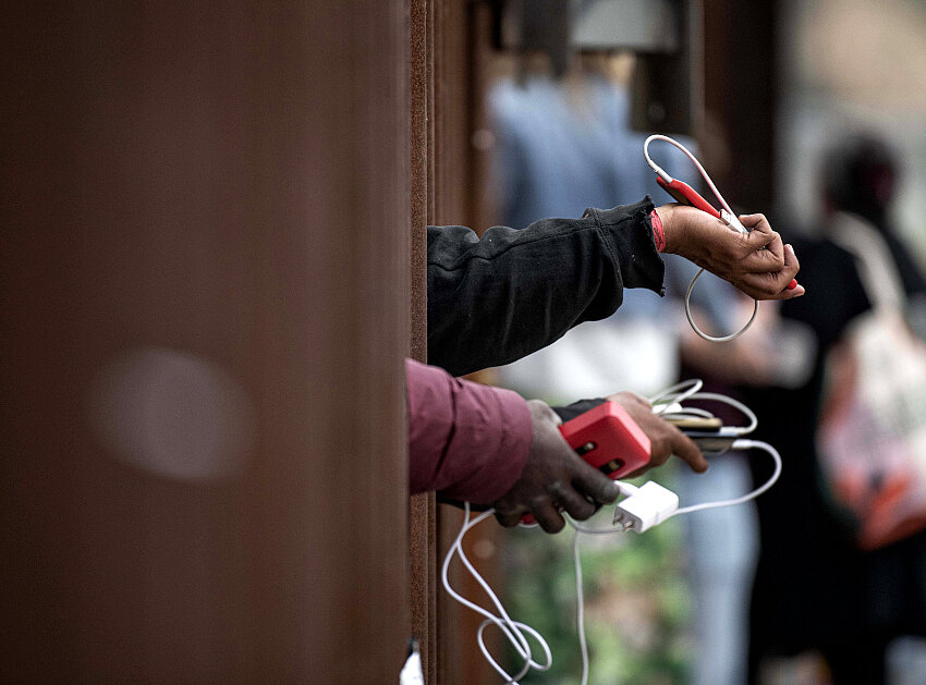 Solicitantes de asilo sostienen sus teléfonos a través del muro fronterizo mientras suplican a la gente que elijan su teléfono para ser cargado