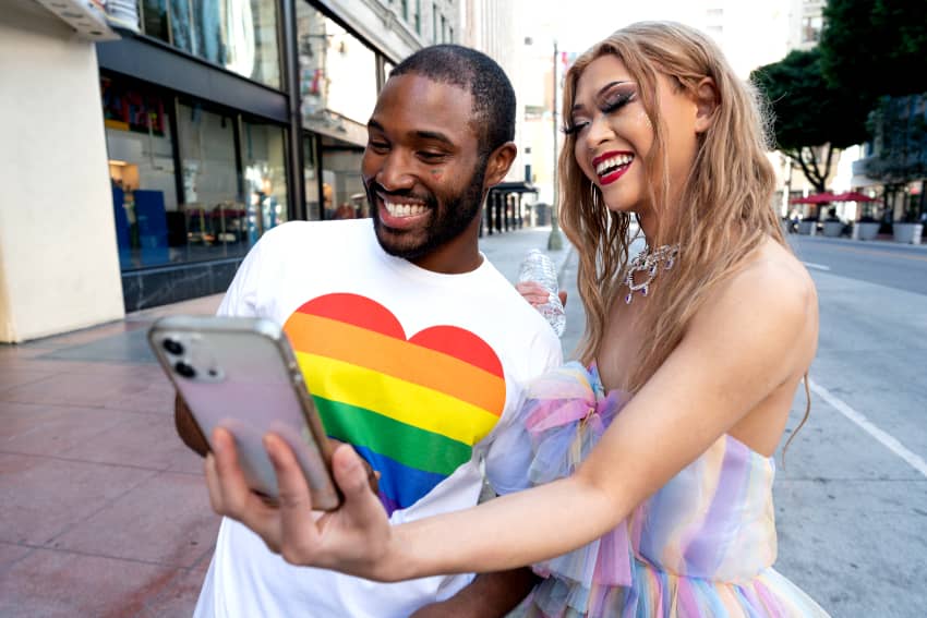 Dos personas se hacen un selfie en la calle; una lleva una camiseta con un corazón arcoíris y la otra un vestido de colores pastel.