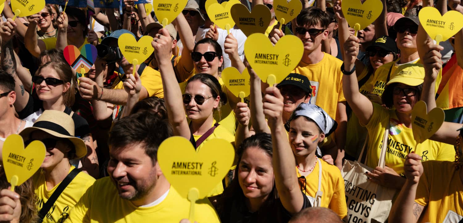 Multitud de personas participantes en una concentración de Amnistía Internacional sostienen corazones amarillos con el logotipo de la organización...