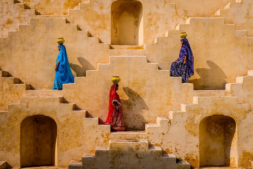 Unas mujeres suben y bajan escalones, mientras llevan ollas de agua en la cabeza.
