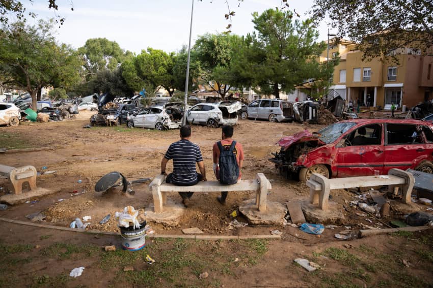 Dos hombres sentados en un banco frente a coches destruidos por las inundaciones en Alfafar tras la DANA de 2024.