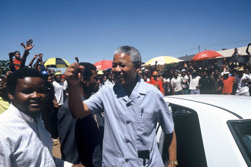 Nelson Mandela levantando el puño y sonriendo ante una multitud en Sudáfrica, símbolo de su liderazgo en la lucha contra el apartheid y el racismo.