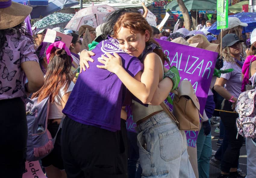 Dos mujeres se abrazan en una marcha feminista, ejemplo de sororidad, apoyo mutuo y lucha colectiva por la igualdad de género y los derechos humanos.