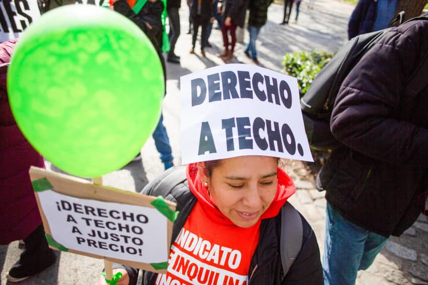 Mujer en una manifestación con un cartel en la cabeza que dice “Derecho a techo” y otro globo verde, rodeada de personas protestando en la calle.