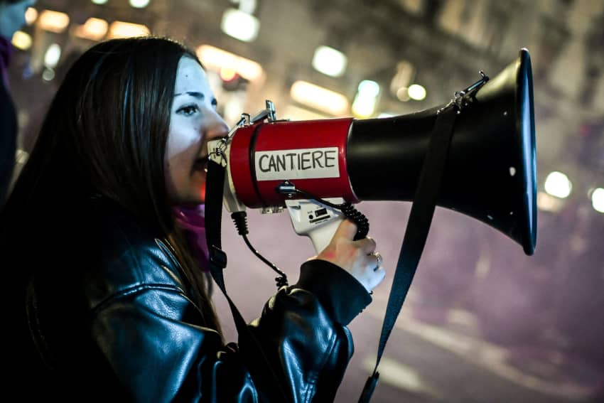 Mujer joven con chaqueta negra habla por un megáfono rojo durante una manifestación nocturna por los derechos de las mujeres