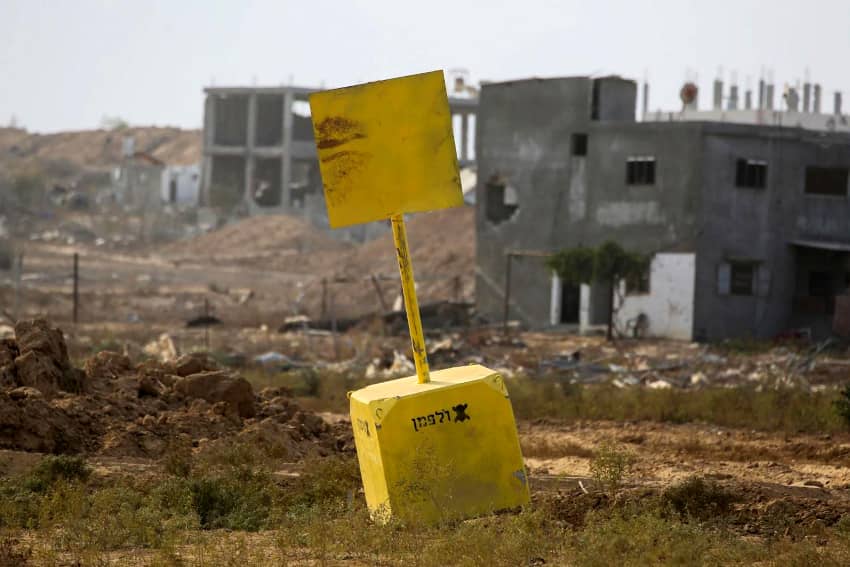 Cubo amarillo de señalización en un terreno devastado de la Franja de Gaza, con edificios dañados al fondo.