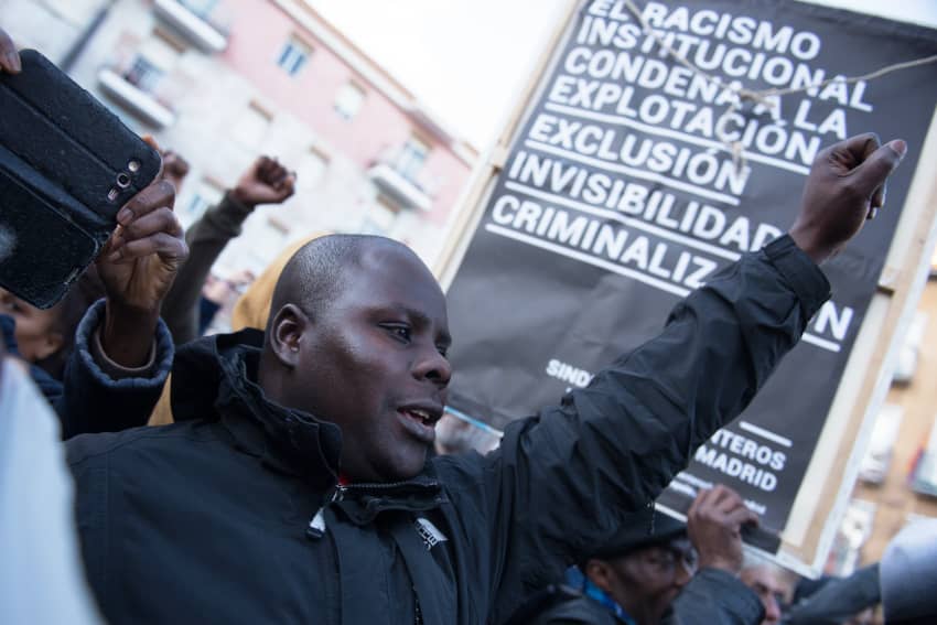 Hombre negro alza el puño durante una protesta en Lavapiés, rodeado de otras personas con el puño en alto y un cartel contra el racismo institucional de fondo.