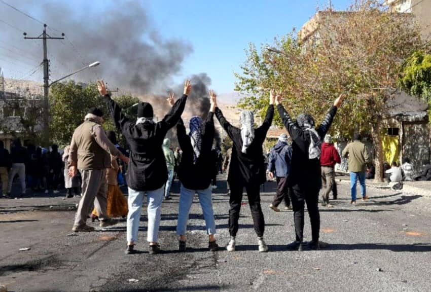 Grupo de personas participa en una protesta en una calle de Mahabad, en la provincia iraní del Kurdistán, mientras se eleva humo al fondo.