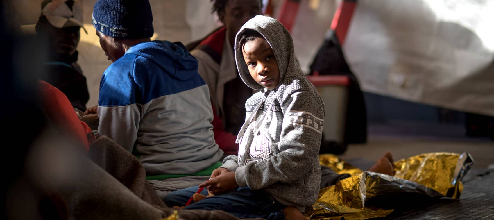 Un niño con una sudadera gris y capucha sentado junto a otras personas rescatadas a bordo del Sea-Watch 3
